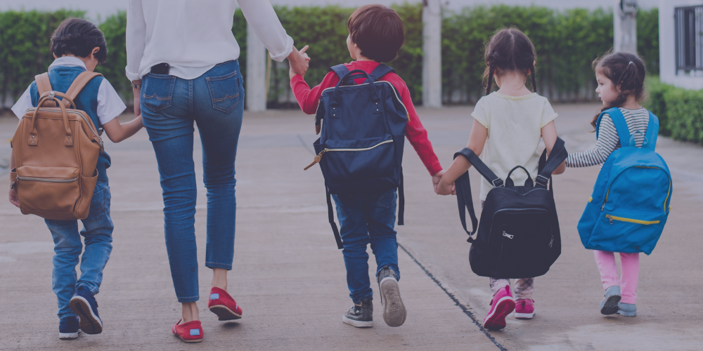 pre-school kids with backpacks walk hand in hand with an adult.